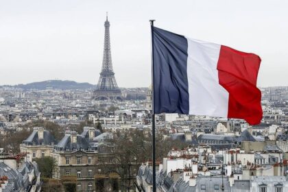 A French Flag Waves Above The Skyline As The Eiffel Tower And Roof Tops Are Seen In Paris, France