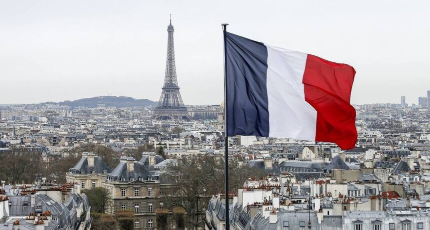 A French Flag Waves Above The Skyline As The Eiffel Tower And Roof Tops Are Seen In Paris, France